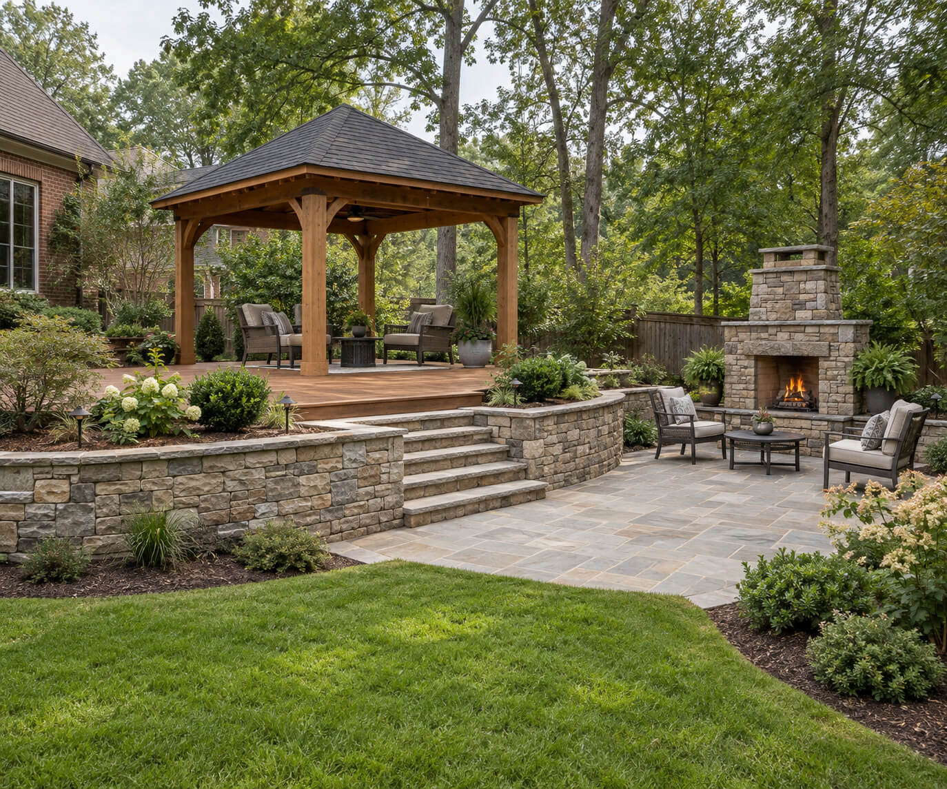 Wooden gazebo on a raised deck with stone retaining walls and steps leading to a patio with an outdoor fireplace.