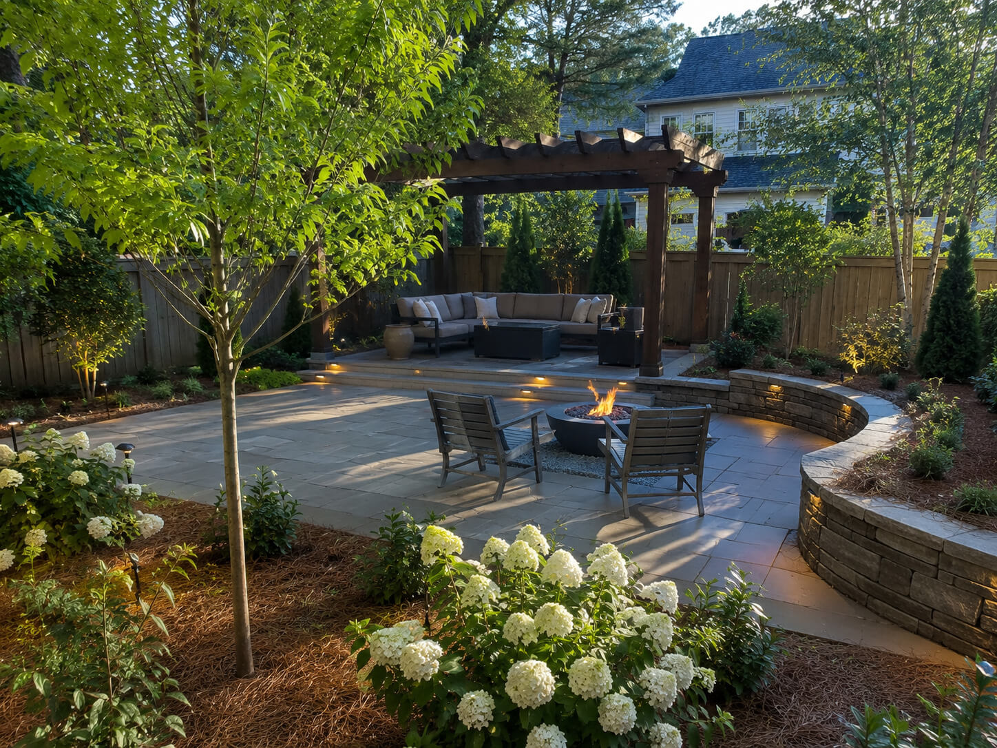 Backyard-Patio-Landscape-with-Pergola Backyard patio landscape with pergola seating area, fire feature, stone patio, landscape lighting, pine straw, and hydrangeas