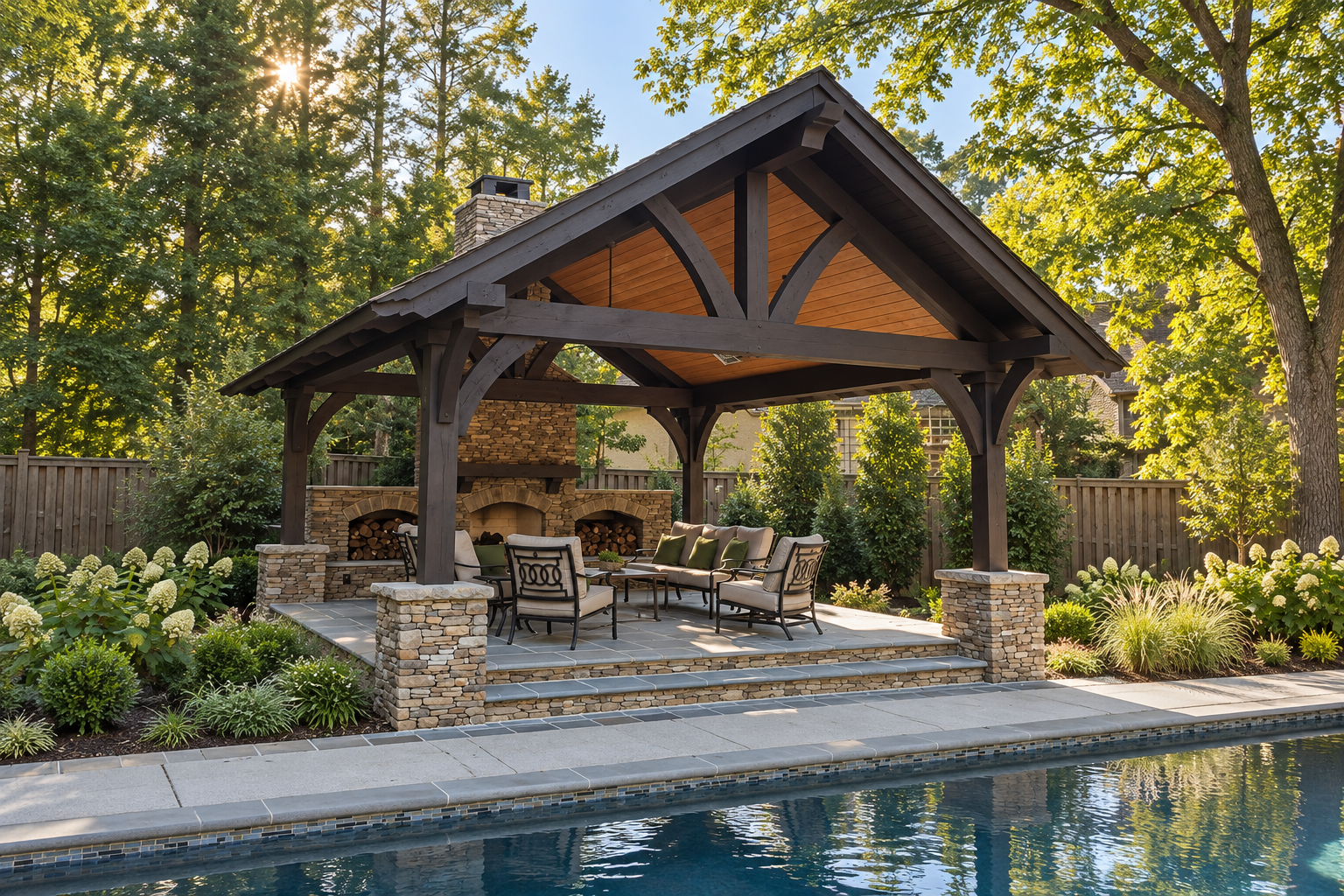 Timber pavilion with a stone fireplace, cushioned seating, and stone patio steps beside a swimming pool.